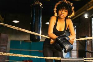 Woman fighter girl power. African american woman fighter with boxing gloves standing on boxing ring leaning on ropes waiting and resting. Strong powerful girl. Strength fit body workout training. photo