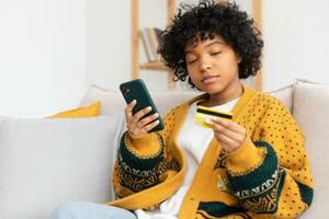 mujer afroamericana comprando en línea con un teléfono inteligente pagando con tarjeta de crédito dorada. niña sentada en casa comprando en internet ingrese los detalles de la tarjeta de crédito. servicio de entrega de comercio electrónico de compras en línea. foto