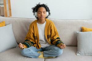 Yoga mindfulness meditation. Young healthy african girl practicing yoga at home. Woman sitting in lotus pose on couch meditating smiling relaxing indoor. Girl doing breathing practice. Yoga at home. photo