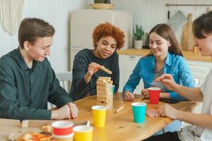 Home party. Friends spending time together playing in board game crash wooden tower at home. Happy diverse group having fun together indoor. Mixed race young buddies best friends enjoying weekend. photo