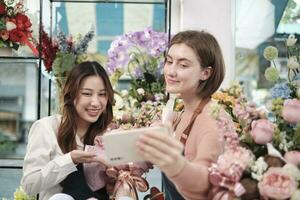 E-commerce business partner, two young female florist workers show floral arrangement, online live streaming, and selfies with smartphone application in bright flower shop, beautiful blossoms store. photo