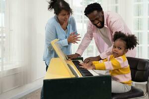 Dad teaching little daughter playing piano at home while mother singing a song. Smiling curly kid looking at camera while sitting with piano photo