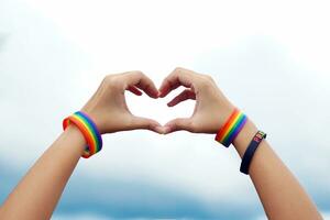 asiático mujer vistiendo arco iris pulseras y negro pulseras impreso con el mensaje orgullo, participación su manos juntos en un corazón forma en contra un cielo antecedentes a espectáculo el lgbt orgullo símbolo. foto