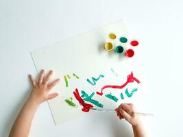 small child draws with paints and brush on white table. photo