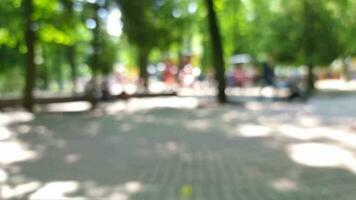 video van een kinderen park met bomen, kinderen en mensen wandelen. de video is wazig en wazig met bokeh. de voorgrond bestaat van kasseien. dag, de weer is zonnig.