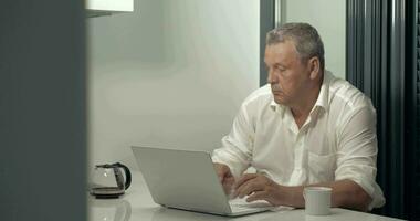 Serious adult man working on laptop while drinking coffee in kitchen video