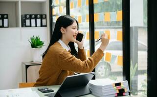 Asian businesswoman holding a pen taking note on note-paper attached to a mirror as a reminder laptop and tablet on the desk.in office photo