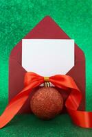 Christmas blank greeting card mockup with red glitter ball with bow and ribbon on sparkling green background. Envelope with letter with white paper. Vertical. Copy space photo