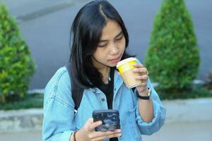 asian digital nomad young woman watching on smartphone seriously for online discussion by video conference and drinking a cup of coffee walking in street, outdoor photo