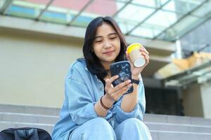 contento asiático joven mujer utilizando solicitud en teléfono inteligente, acecho video, surf en social medios de comunicación redes y bebida un taza de café mientras sentado en un centro comercial escalera foto