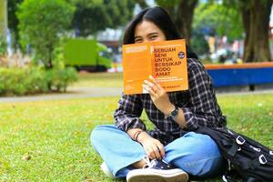 happy young asian woman college student cover up her face with a book looking to camera photo