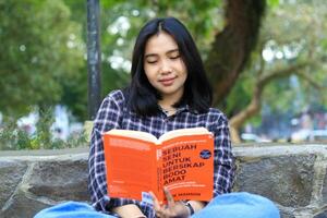contento consciente de joven asiático mujer Universidad estudiante leyendo un libro en el parque, educación concepto foto