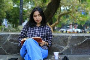 contento consciente de joven asiático mujer Universidad estudiante leyendo un libro en el parque, educación concepto foto