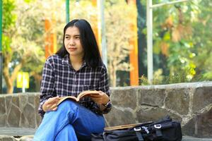 happy young asian college student read a book in the park looking away photo