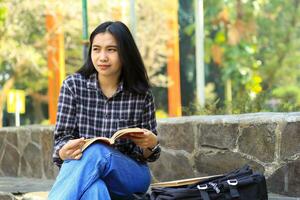 happy young asian college student read a book in the park looking away photo