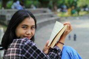 contento joven asiático mujer Universidad estudiante con sonriente cara disfrutar leer un libro para examen en el parque foto