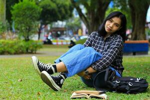 peaceful asian college student smiling and sitting in the lawn with book and backpack photo