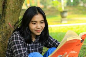 contento y éxito hembra asiático Universidad estudiante disfrutando leer un libro en el parque foto
