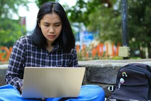 happy young asian woman focused using laptop  working remotely and browsing in social media in comfortable outdoors space photo