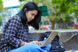 happy young asian woman focused using laptop  working remotely and browsing in social media in comfortable outdoors space photo