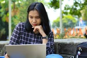 happy young asian woman focused using laptop  working remotely and browsing in social media in comfortable outdoors space photo