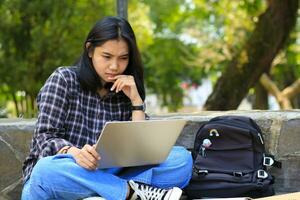 happy young asian woman focused using laptop  working remotely and browsing in social media in comfortable outdoors space photo