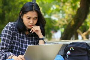 happy young asian woman focused using laptop  working remotely and browsing in social media in comfortable outdoors space photo