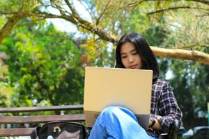 happy young asian woman focused using laptop  working remotely and browsing in social media in comfortable outdoors space photo