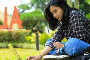 happy young asian woman focused using laptop  working remotely and browsing in social media in comfortable outdoors space photo