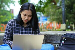 happy young asian woman focused using laptop  working remotely and browsing in social media in comfortable outdoors space photo