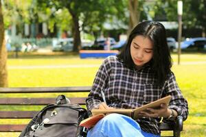 beautiful asian young woman college student focused writing on notebook and reading book in outdoors city park photo