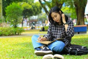 beautiful asian young woman college student focused writing on notebook and reading book in outdoors city park photo