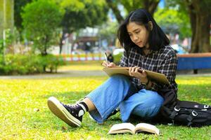 beautiful asian young woman college student focused writing on notebook and reading book in outdoors city park photo