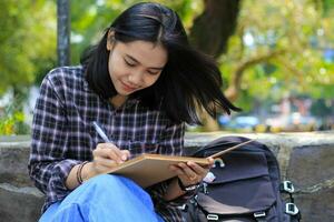 sonriente asiático hermosa joven mujer disfrutar escritura a hacer lista y idea en cuaderno en al aire libre ciudad parque foto