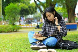 hermosa asiático joven mujer Universidad estudiante enfocado escritura en cuaderno y leyendo libro en al aire libre ciudad parque foto