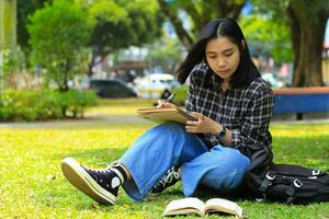 hermosa asiático joven mujer Universidad estudiante enfocado escritura en cuaderno y leyendo libro en al aire libre ciudad parque foto