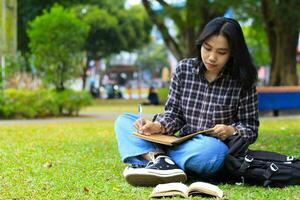 sonriente asiático hermosa joven mujer disfrutar escritura a hacer lista y idea en cuaderno en al aire libre ciudad parque foto