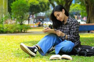 sonriente asiático hermosa joven mujer disfrutar escritura a hacer lista y idea en cuaderno en al aire libre ciudad parque foto