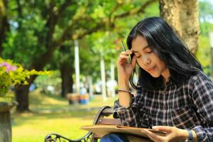 confusing asian young woman thinking an idea and writing in notebook with a pen in nature outdoors area photo