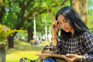 confusing asian young woman thinking an idea and writing in notebook with a pen in nature outdoors area photo