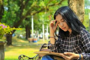 confusing asian young woman thinking an idea and writing in notebook with a pen in nature outdoors area photo