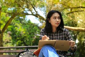 portrait of relax young asian woman writing idea and memories on notebook im outdoors area or park look away photo