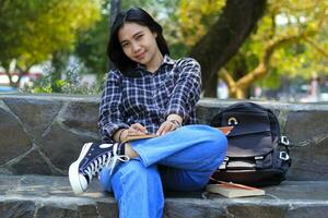 smiling attractive asian young woman writing to do list and organizing her job on notebook in city park photo