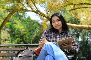 smiling attractive asian young woman writing to do list and organizing her job on notebook in city park photo