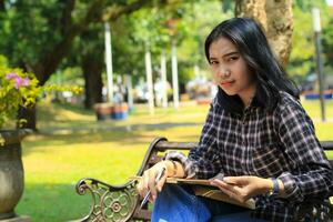 smiling attractive asian young woman writing to do list and organizing her job on notebook in city park photo