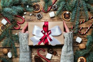 Female hands hold Gift box with bow. Christmas tree and decorations, cane caramel and oranges nuts Wooden background photo