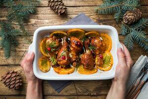 Baked chicken drumstick with oranges and cranberries in a baking sheet holding women's hands on a wooden background. Christmas food Table with decorations. photo