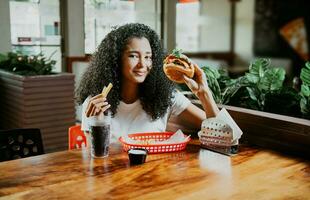 Happy girl enjoying a delicious hamburger sitting in a restaurant. Portrait of young afro woman holding a hamburger with fries in a restaurant photo
