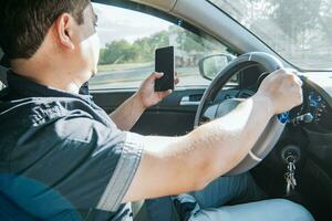Person holding the cell phone and with the other hand the steering wheel, Man using his phone while driving, Concept of irresponsible driving, Distracted driver using the cell phone while driving photo