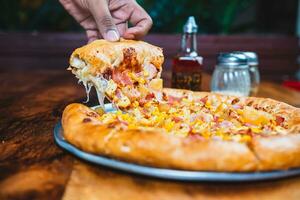 Homemade ham pizza with cheese and sweet corn served on wooden table. Hand taking a slice of ham and sweetcorn pizza photo